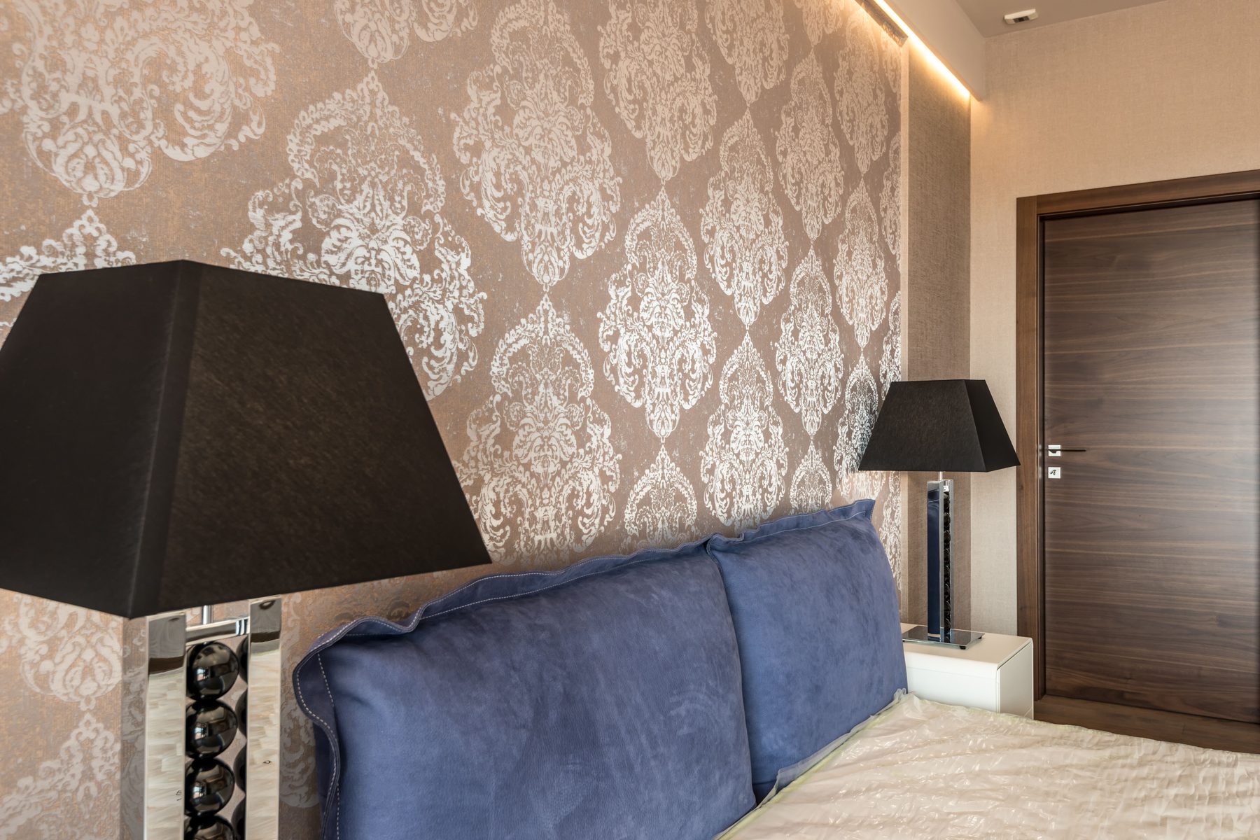 Bedroom wall covered in a metallic bronze damask wallpaper behind a bed with blue velvet pillows and black bedside lamps