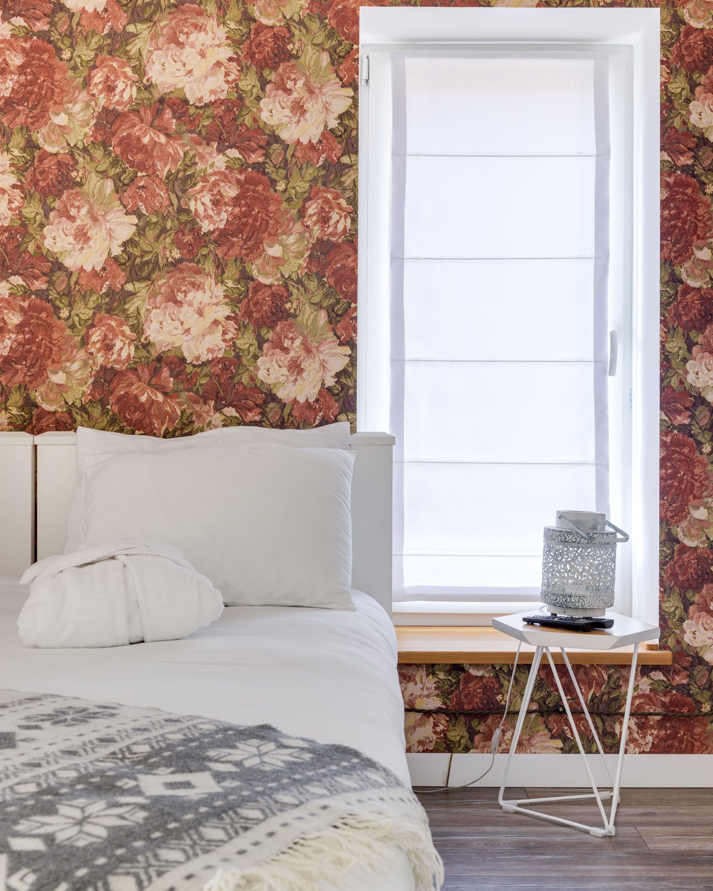 Bedroom with a bold red and pink rose wallpaper, a white bed with a grey knit throw, and a window with a roman blind